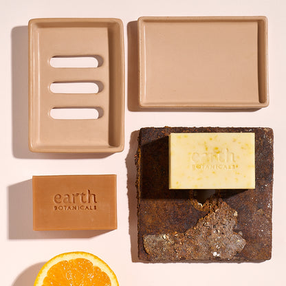 Two soap bars from 'earth BOTANICALS' on a textured surface with a Jesmonite Terracotta tray and an orange slice.